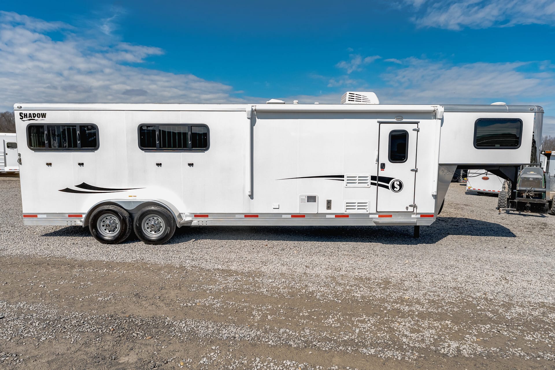 White Shadow travel trailer parked on a gravel lot under a blue sky, showing multiple windows and wheels along its side.