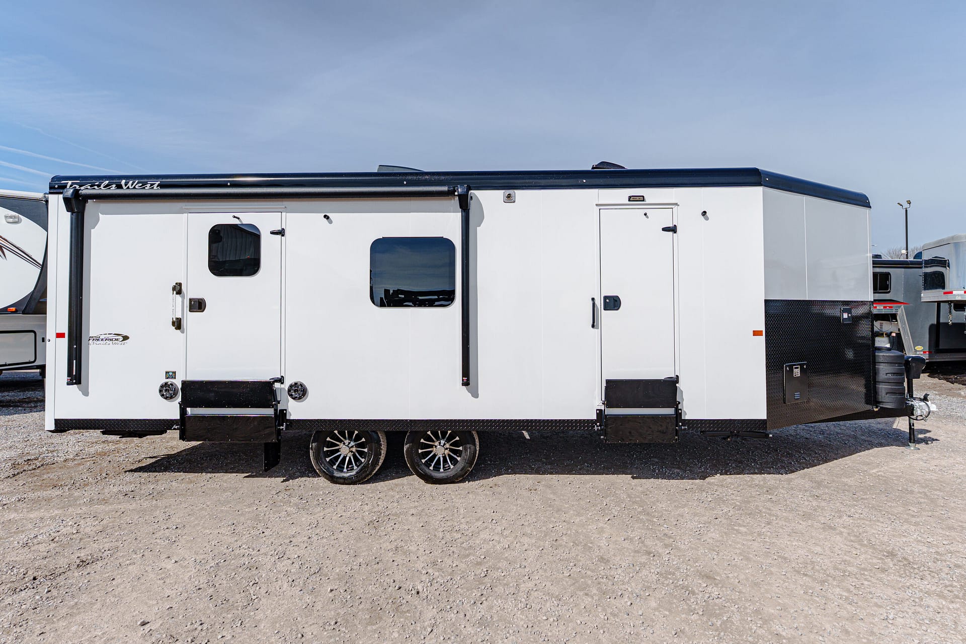 White travel trailer with black trim and two doors parked on a gravel lot under a clear blue sky. It has several small windows and tandem wheels visible.