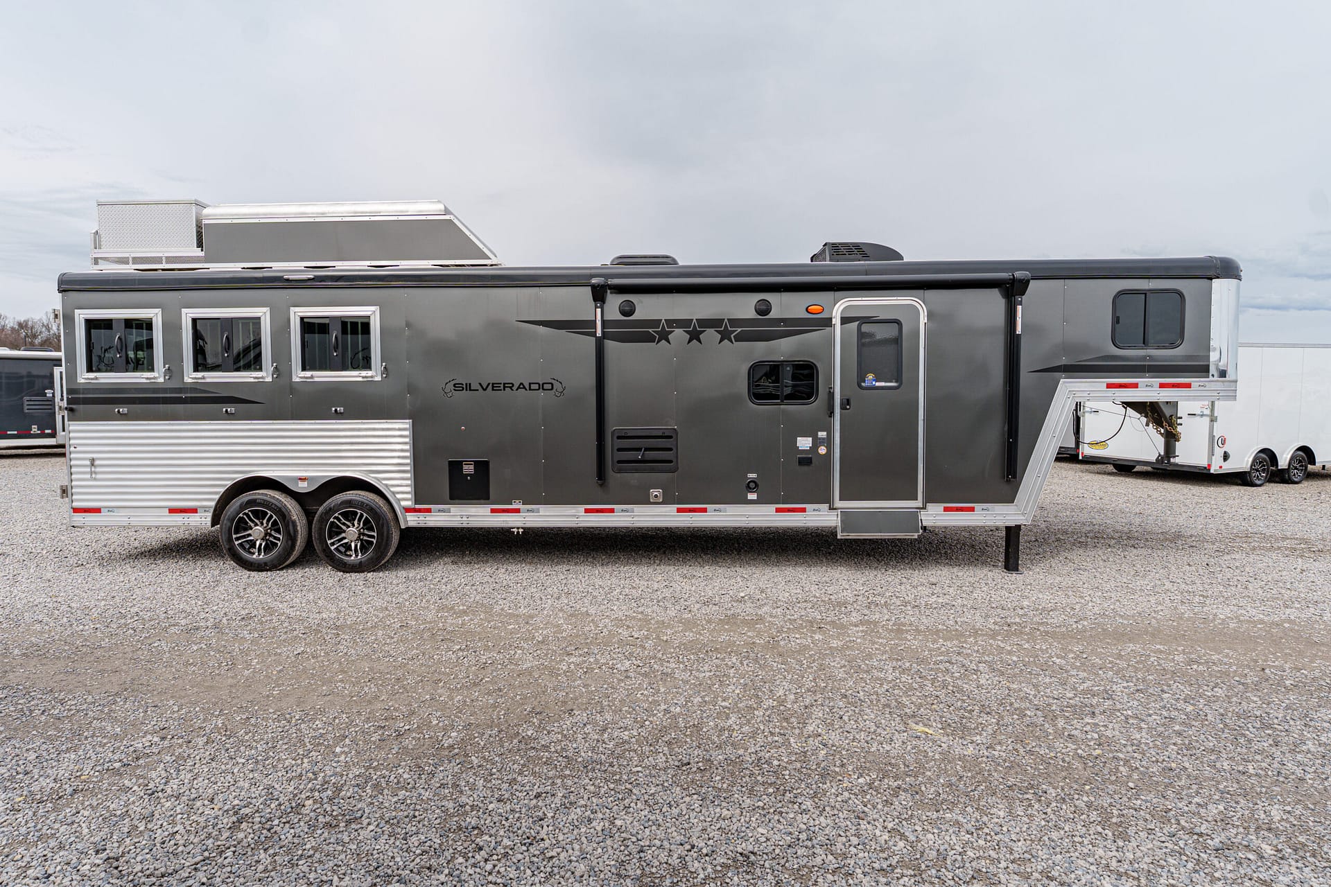 Large gray two-axle travel trailer with star graphics, parked on a gravel lot under a cloudy sky, side door and windows visible.