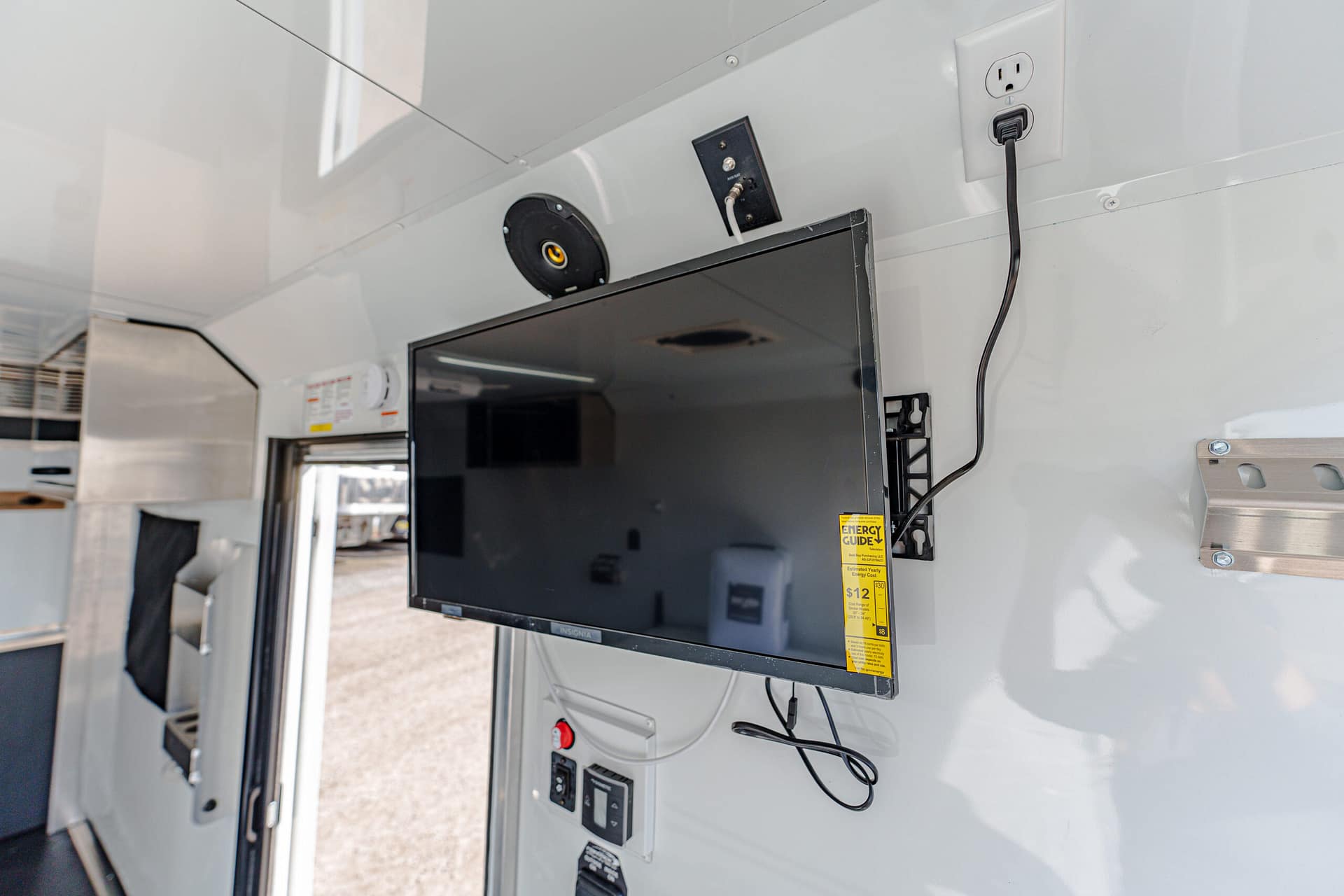 Flat-screen TV mounted on a gray wall inside a vehicle or trailer, with electrical outlets and cables nearby.