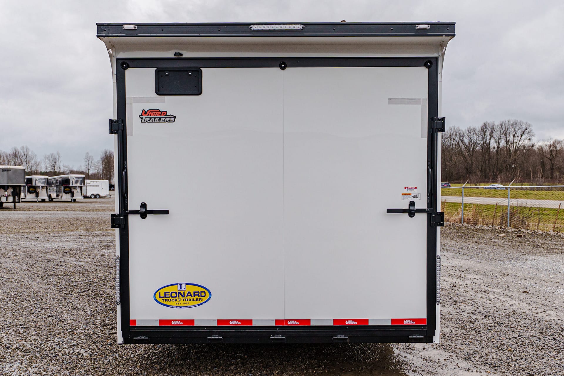 Rear of a white trailer with a Leonard Truck & Trailer logo, parked on a gravel lot with other trailers in the distance under a cloudy sky.