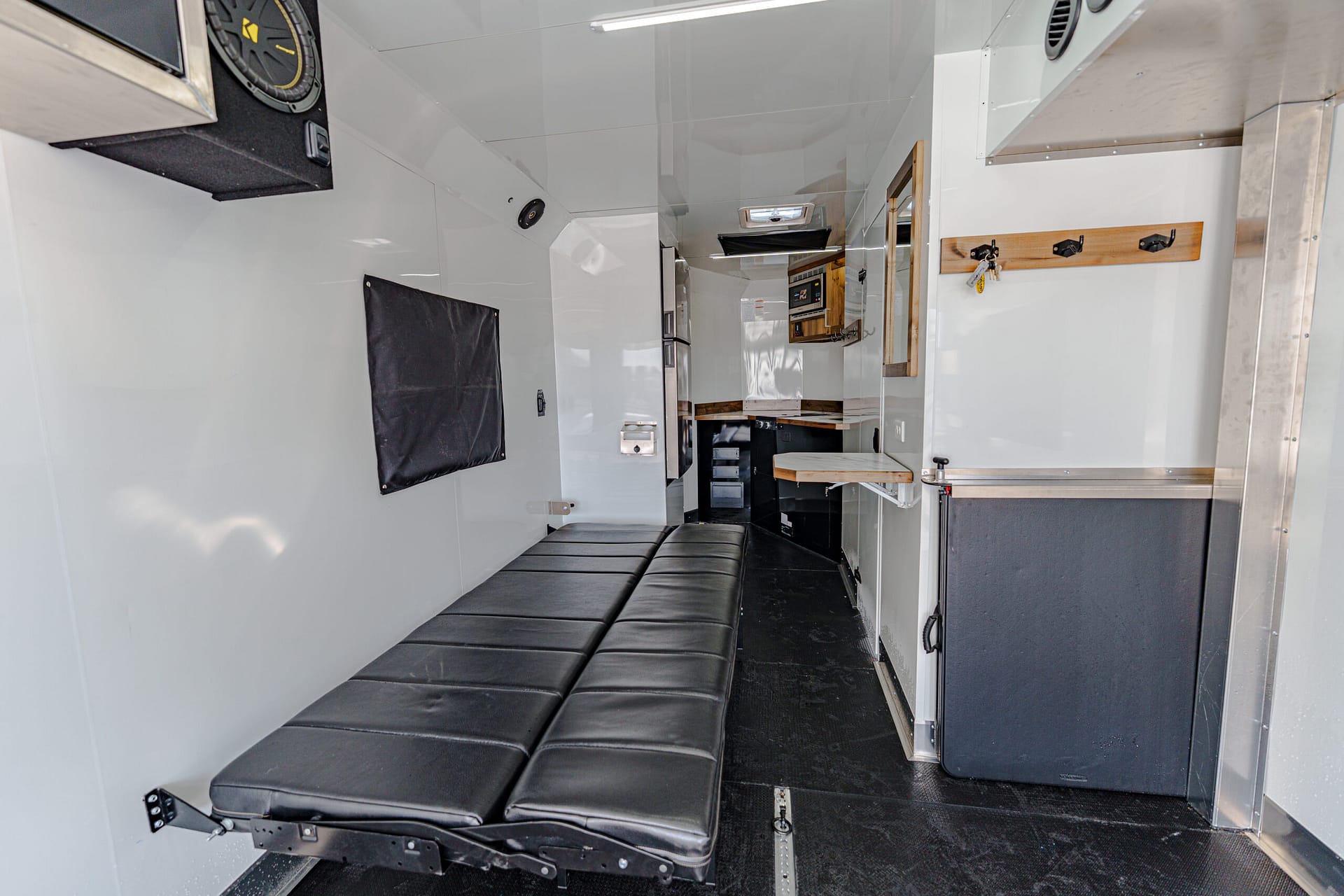 Interior of a white mobile workspace/trailer with a long black padded bench on the left and wooden hooks on the right wall.