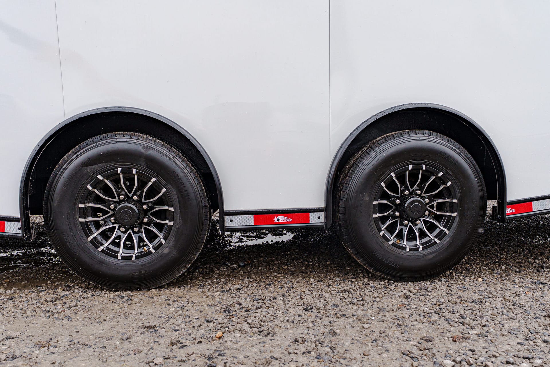 Two black trailer wheels with silver rims under a pale gray trailer body, shown side by side.