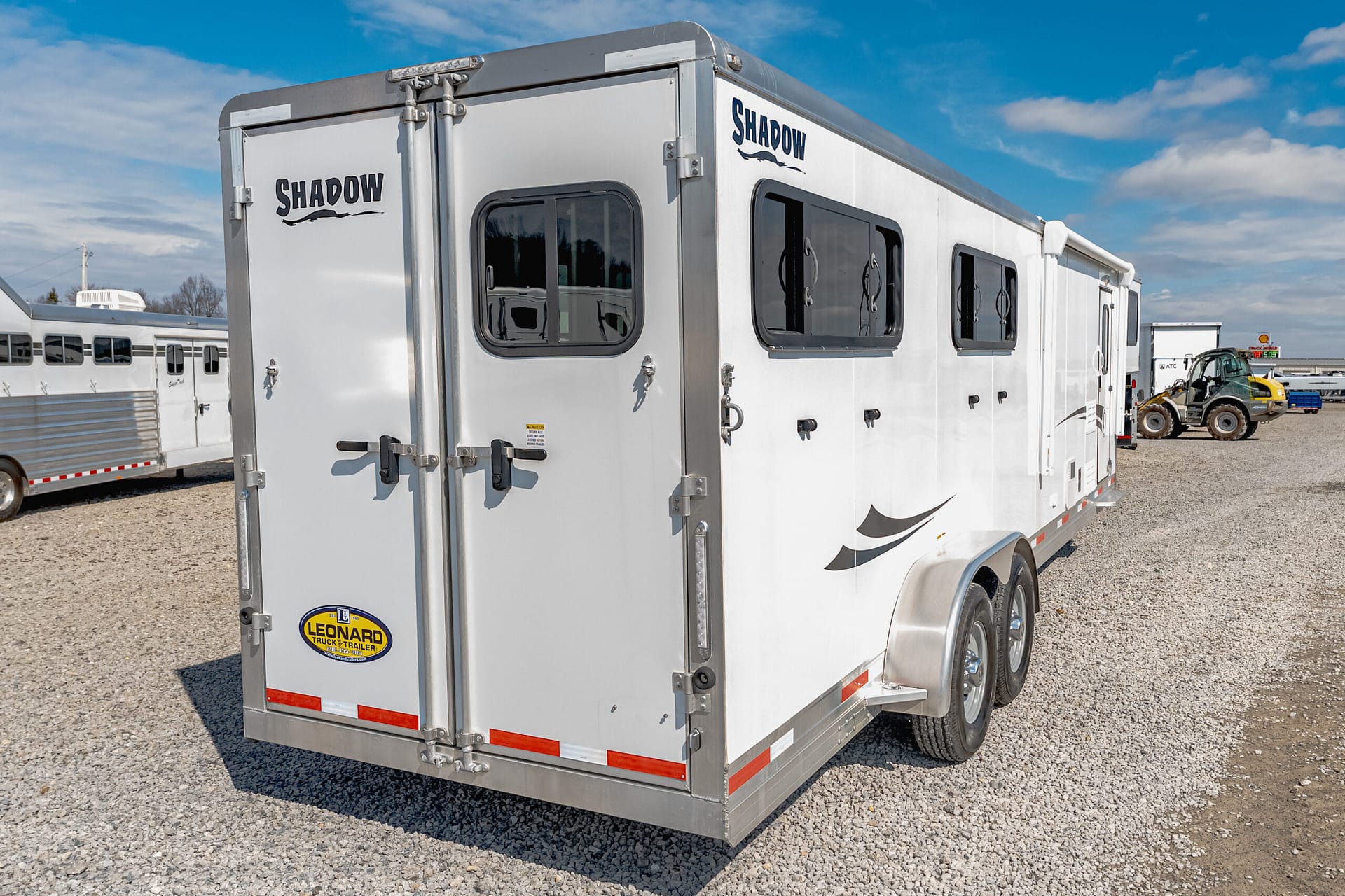 White Shadow horse trailer with two rear doors and side windows parked in a gravel lot under a blue sky.