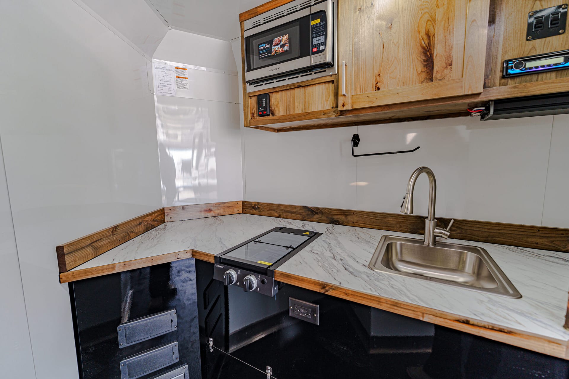 Compact RV kitchen: microwave above wooden cabinets, marble countertop, and a stainless sink with faucet on the right.