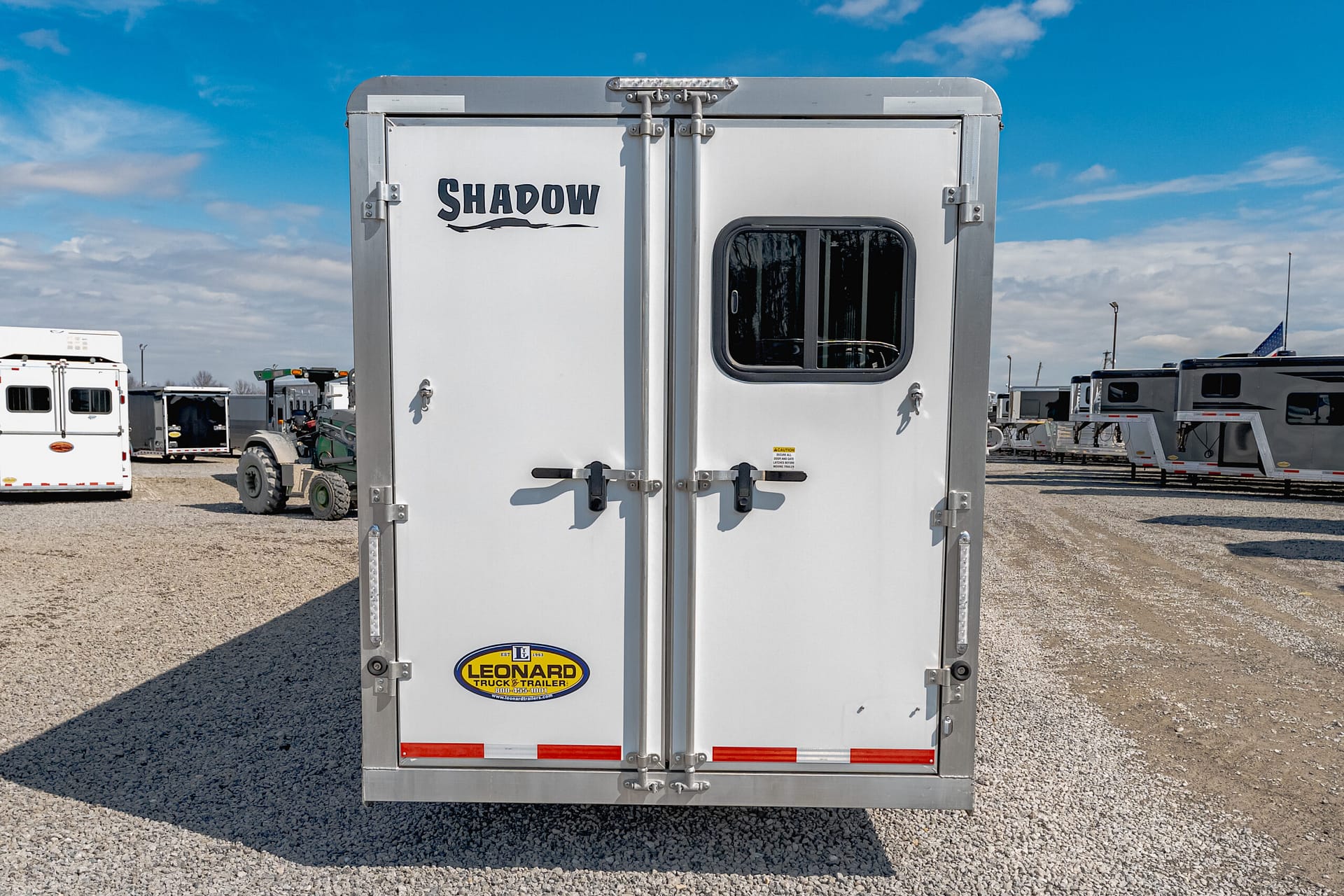 Rear view of a silver trailer with a small rounded window and Leonard Truck & Trailer logo, parked in a gravel lot with other trailers nearby.