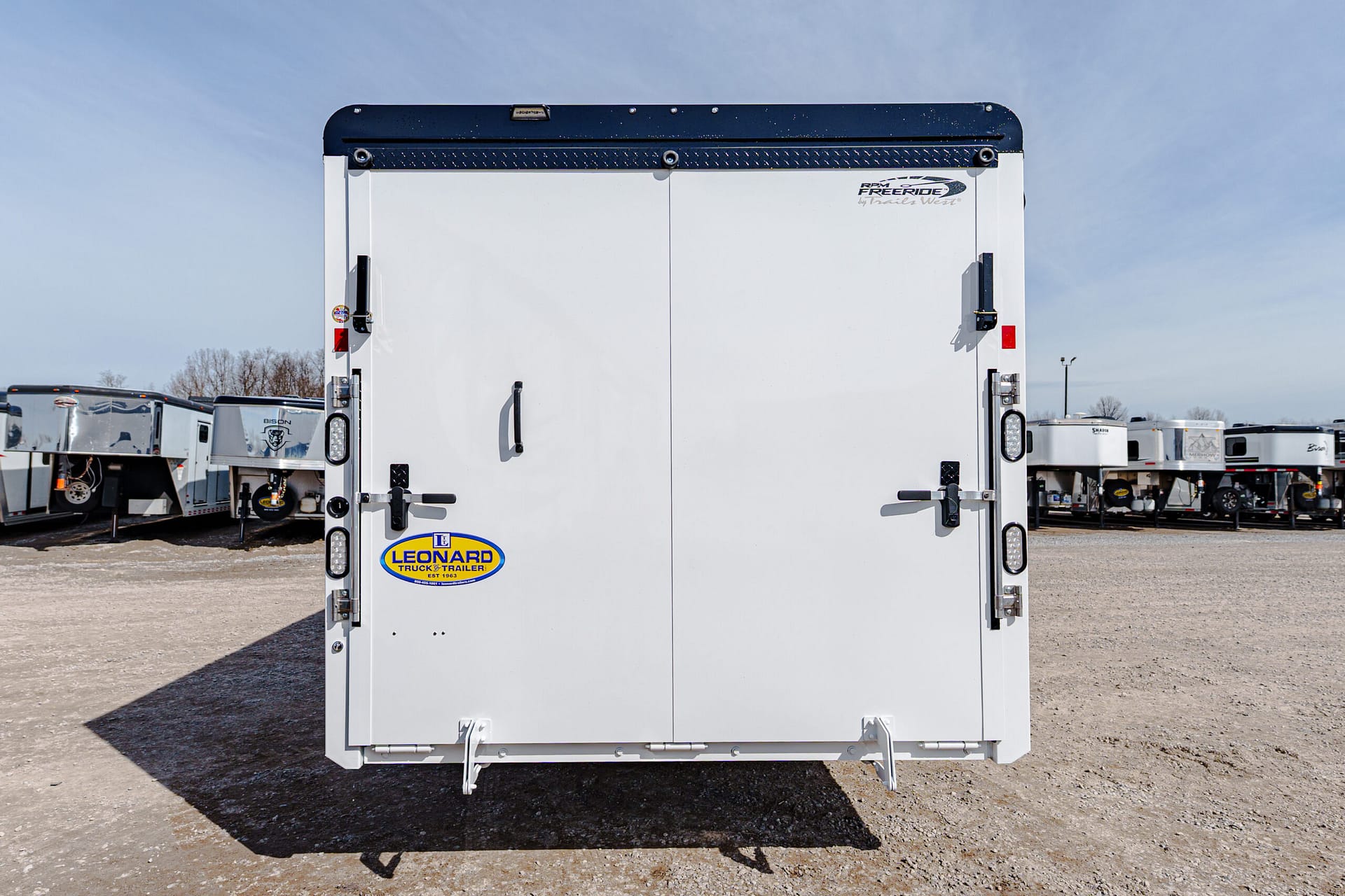 Rear view of a white cargo trailer in a lot, doors closed, with Leonard Truck & Trailer logo on the lower left door and other trailers in the background.
