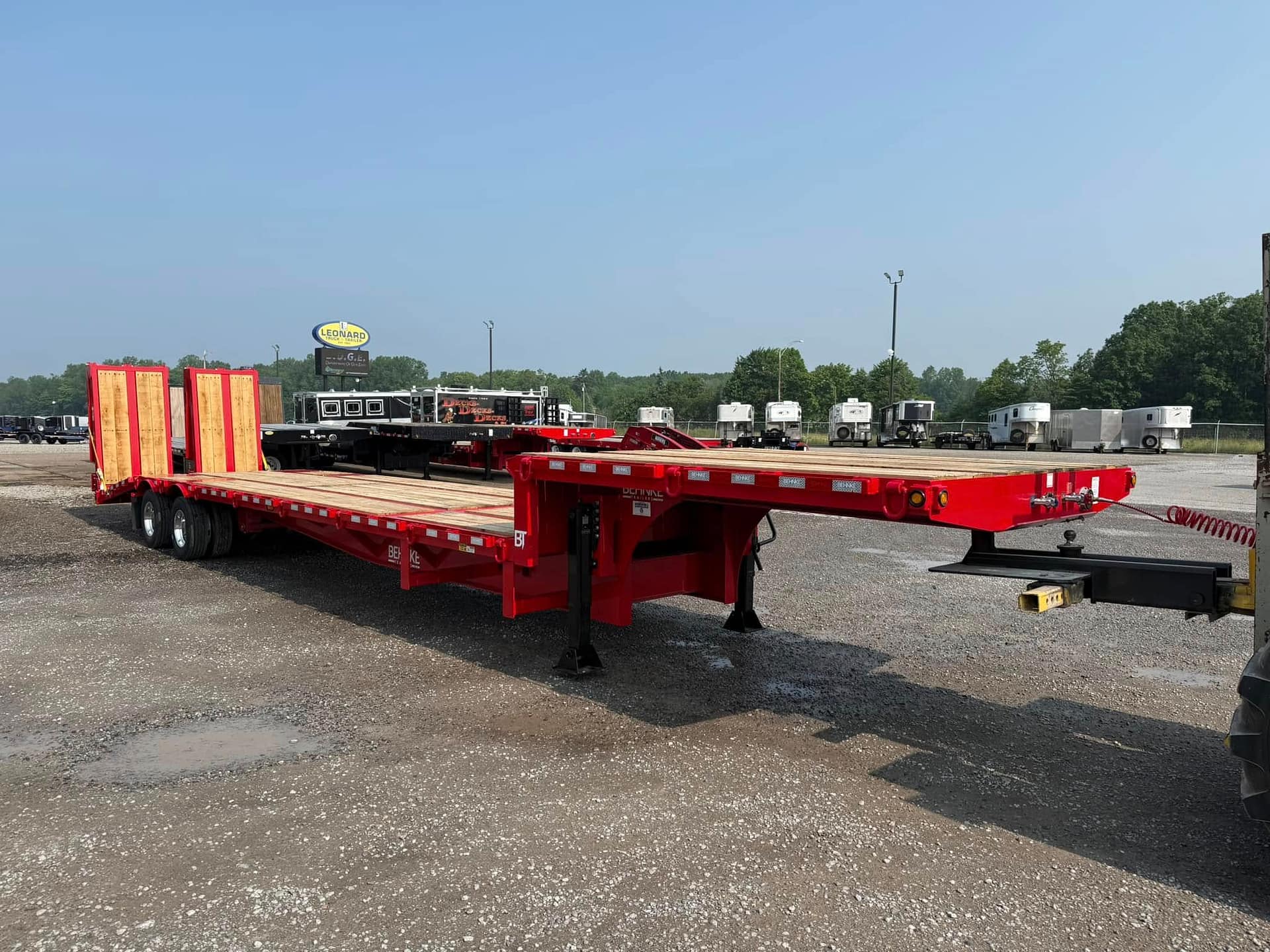 Red flatbed trailers parked in a lot with white horse trailers in the background under a clear blue sky, with a Leonard sign visible in the distance.