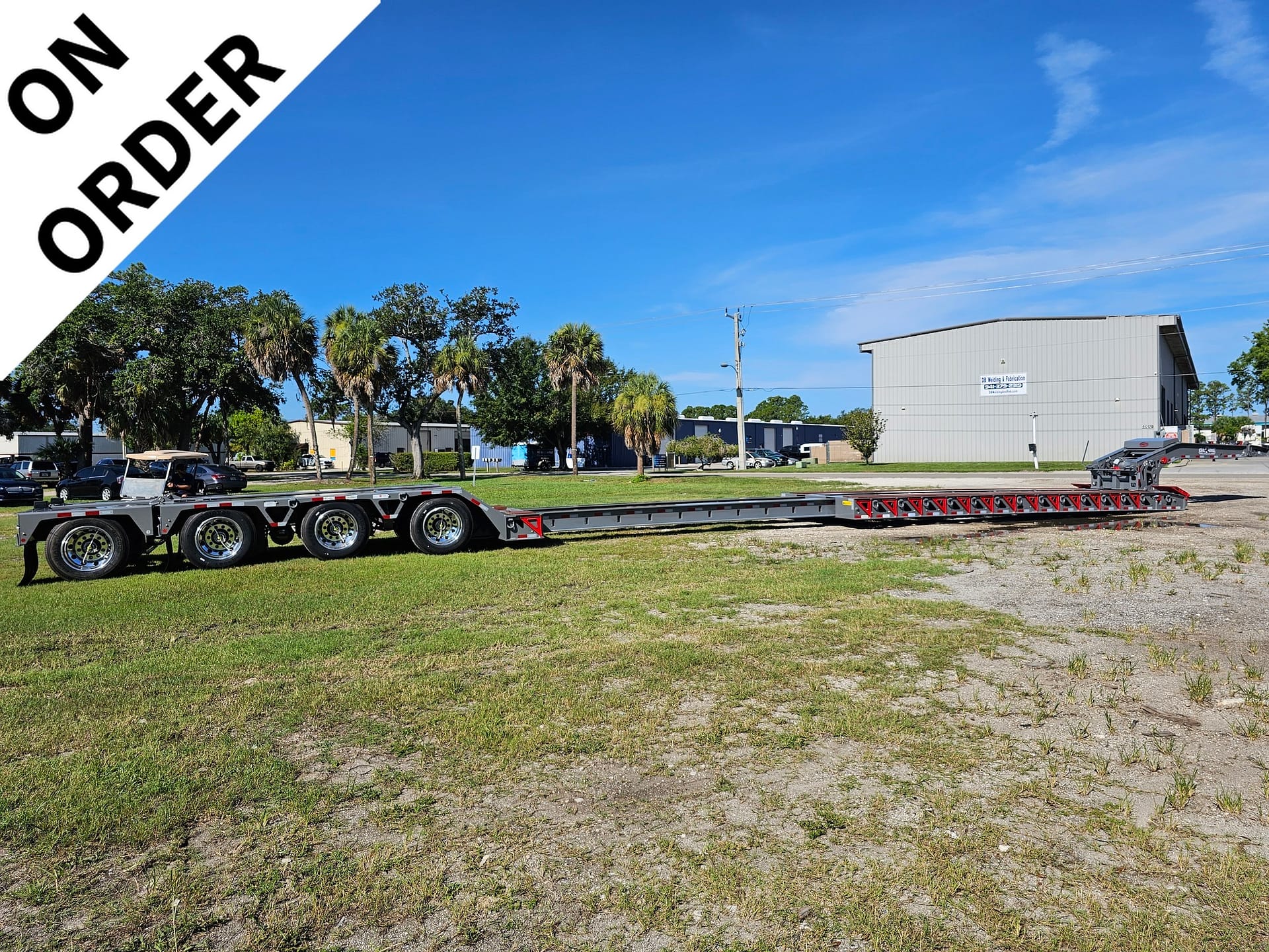 Industrial parking lot with a long multi-axle trailer in foreground, gray warehouse on the right, palm trees, and a diagonal white banner reading 'ONE ORDER' across the top-left.