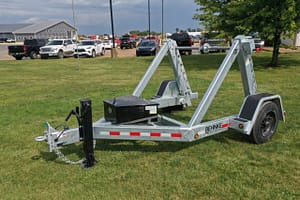 DJI_0129-scaled – Leonard Truck and Trailer, Inc Galvanized boat trailer on a grassy field with a line of parked cars and a gray industrial building in the background.