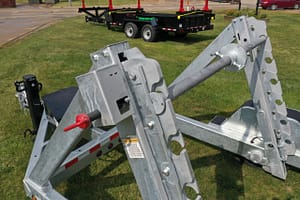 DJI_0137-scaled – Leonard Truck and Trailer, Inc Close-up of a galvanized steel industrial frame with bolts and red handles on grass; a flatbed trailer sits in the background.