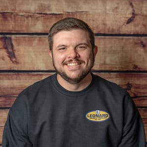 Smiling man with short hair and beard wearing a black sweatshirt with a Leonard Truck & Trailer logo, wooden backdrop.