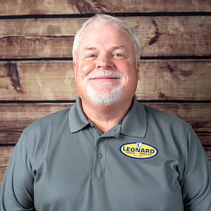 Smiling older man with gray hair and a white beard wearing a Leonard Truck & Trailer polo, standing against a wooden wall.