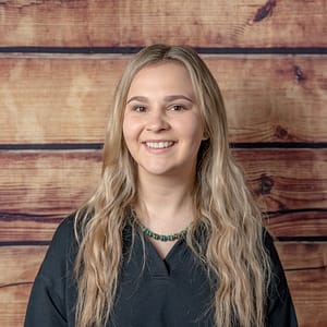 Smiling young woman with long wavy blonde hair wearing a teal bead necklace and a black top, standing against a wooden background.