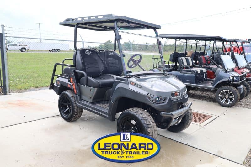 Gray off-road golf cart with a roof, two seats, and a cargo bed on a dealership lot, with more carts in the background and the Leonard Truck & Trailer sign in view.