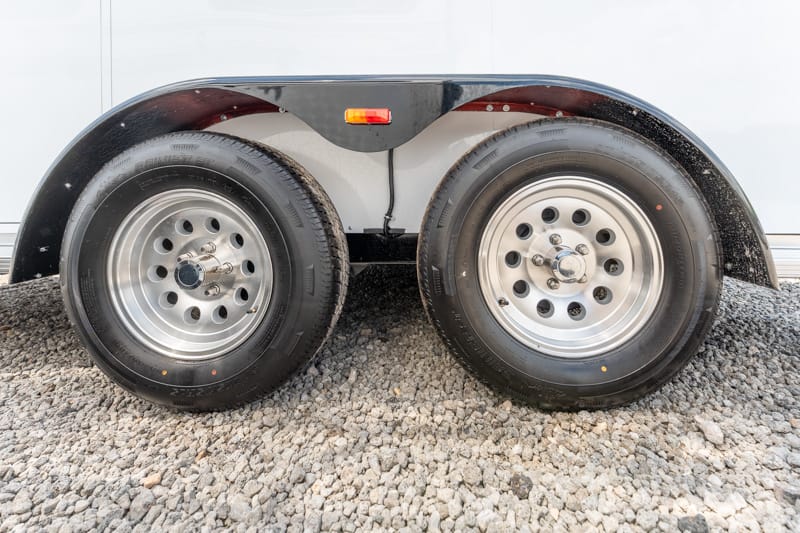 Dual trailer wheels with silver rims on gravel under a trailer fender, showing the tandem axle. **(informative)**