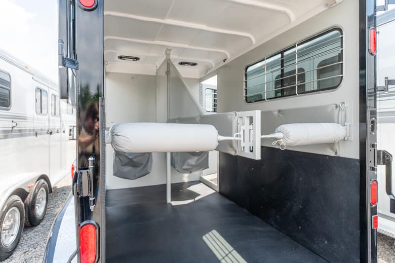 Interior of a horse trailer showing two padded stalls and divider bars for transporting horses, with a rubberized floor and side windowed wall.