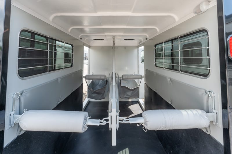 Interior of a two-horse trailer with separated stalls, padded walls, and barred windows on each side under a white ceiling.