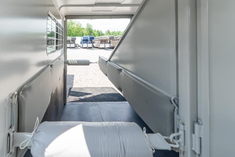 Interior of a light-gray horse trailer with padded walls and a central aisle, open to a parking lot with other trailers outside.