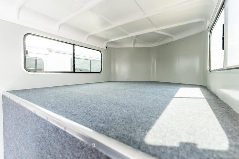 Interior of a small white trailer with a gray carpeted bed/platform and side windows along the walls.