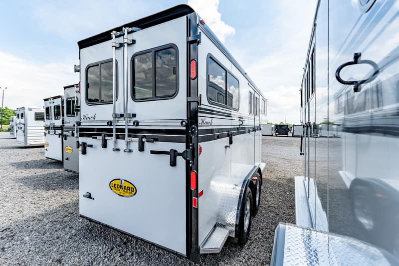 White tandem-axle travel trailer with Leonard branding parked in a gravel lot, reflection visible on neighboring trailer side, with more trailers in the background.