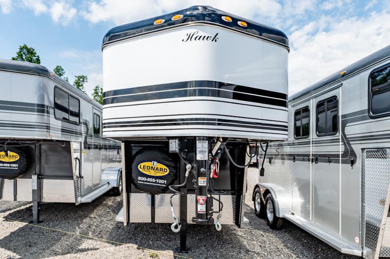 Front view of a white Hawk fifth-wheel trailer on a gravel lot, flanked by two other RVs under a blue sky with scattered clouds.
