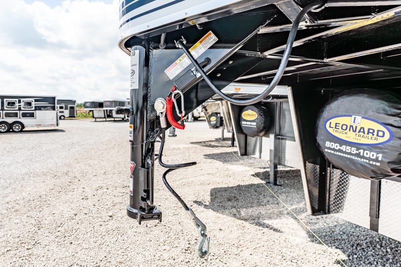 Close-up of a trailer hitch and jack under a Leonard trailer, with safety chains and wiring visible in a gravel lot.