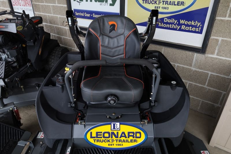 Zero-turn lawn mower cockpit with black seat and orange stitching, joystick arms, and Leonard Truck & Trailer sign in background.