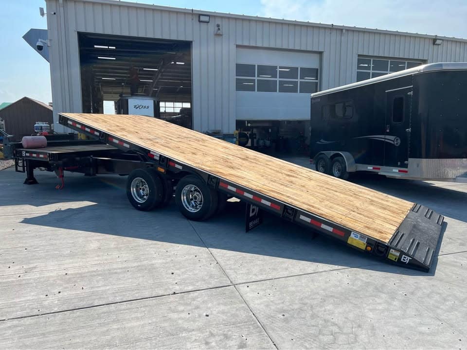 Flatbed trailer with a wooden ramp extended at a repair shop near a black enclosed trailer and a warehouse building in the background