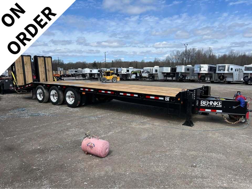 Long Behnke flatbed trailer with wooden deck and three axles in a yard among other trailers, with an 'ON ORDER' banner.